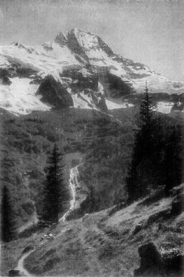 A photograph of a snow-capped mountain near the source of the Rhine, with a small waterfall and trees in the valley below it.