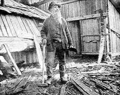 Old miner with long white beard standing in shingle scraps, two right-angle shingle shacks.