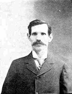 Broad moustache; patterned half-Windsor tie, top-buttoned small-woven-check jacket.