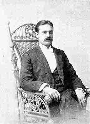 White bow tie, dark three-piece double-breasted suit; seated in ornate bentwood and cane chair.