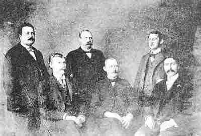 Six men, three sitting, three standing behind, in variety of previously described men's dress.
